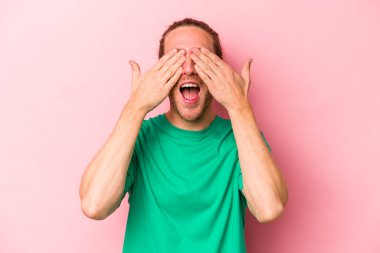 Pembe arka planda soyutlanmış genç beyaz adam elleriyle gözlerini kapatıyor, geniş bir gülümsemeyle bir sürpriz bekliyor..