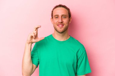 Pembe arka planda izole edilmiş genç beyaz adam işaret parmaklarıyla küçük bir şey tutuyor, gülümsüyor ve kendinden emin..