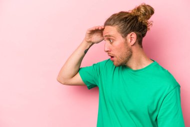 Pembe arka planda izole edilmiş genç beyaz adam uzaklara bakıyor ve elini alnına koyuyor..