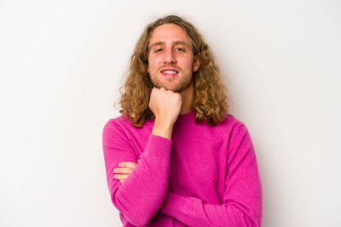 Beyaz arka planda izole olmuş genç beyaz bir adam gülümsüyor ve kendinden emin, çenesine elleriyle dokunuyor..