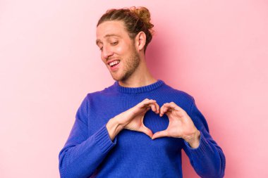 Pembe arka planda izole edilmiş genç beyaz adam gülümsüyor ve elleriyle kalp şeklini gösteriyor..