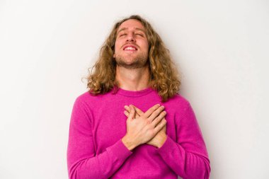 Beyaz arka planda soyutlanmış beyaz tenli genç bir adam gülüyor, elini yüreğine koyuyor, mutluluk kavramını koruyor..