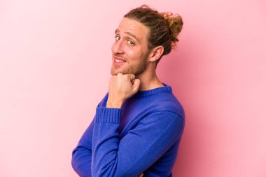 Pembe arka planda izole edilmiş genç beyaz adam gülümsüyor ve kendinden emin, elleriyle çenesine dokunuyor..