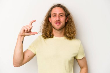 Beyaz arka planda izole edilmiş genç beyaz bir adam işaret parmaklarıyla küçük bir şey tutuyor, gülümsüyor ve kendinden emin..