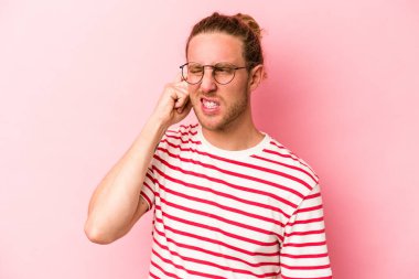 Pembe arka planda izole edilmiş genç beyaz bir adam parmaklarıyla kulaklarını kapatıyor, gürültülü bir ortam yüzünden stresli ve çaresiz..