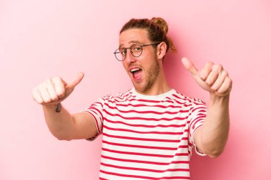Pembe arka planda izole edilmiş genç beyaz adam iki parmağını da kaldırıyor, gülümsüyor ve kendinden emin..