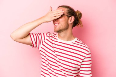 Pembe arka planda soyutlanmış genç beyaz adam elleriyle gözlerini kapatıyor, geniş bir gülümsemeyle bir sürpriz bekliyor..