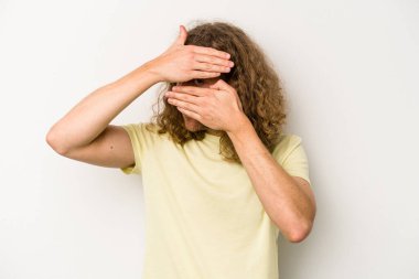 Beyaz arka planda izole edilmiş genç beyaz bir adam kameraya parmaklarıyla göz kırpıyor, yüzünü kapatmaktan utanıyor..