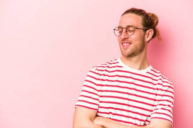 Pembe arka planda izole edilmiş genç beyaz adam kollarını kavuşturarak gülümsüyor..