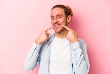 Pembe arka planda izole edilmiş genç beyaz adam gülümsüyor, parmaklarını ağıza doğrultuyor..