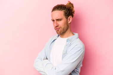 Pembe arka planda izole edilmiş genç beyaz adam hoşnutsuzca kaşlarını çatmış, kollarını kavuşturmuş..