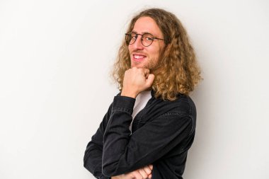 Beyaz arka planda izole olmuş genç beyaz bir adam gülümsüyor ve kendinden emin, çenesine elleriyle dokunuyor..