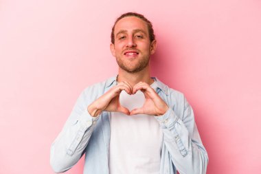 Pembe arka planda izole edilmiş genç beyaz adam gülümsüyor ve elleriyle kalp şeklini gösteriyor..