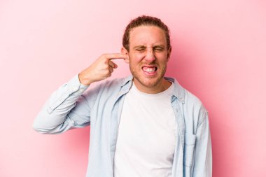 Pembe arka planda izole edilmiş genç beyaz adam kulaklarını elleriyle kapatıyor..