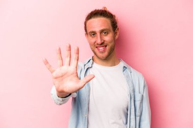 Pembe arka planda izole edilmiş genç beyaz adam gülümseyerek beş numarayı parmaklarıyla gösteriyor..