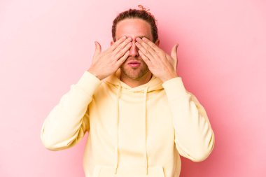 Pembe arka planda izole edilmiş genç beyaz adam elleriyle gözlerini kapatmaktan korkuyor..