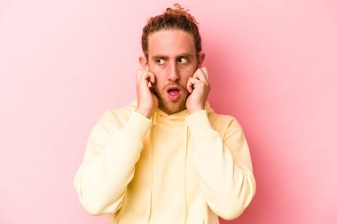 Pembe arka planda izole edilmiş genç beyaz bir adam parmaklarıyla kulaklarını kapatıyor, gürültülü bir ortam yüzünden stresli ve çaresiz..