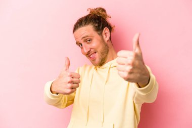 Pembe arka planda izole edilmiş genç beyaz adam iki parmağını da kaldırıyor, gülümsüyor ve kendinden emin..