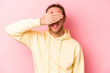 Pembe arka planda soyutlanmış genç beyaz adam elleriyle gözlerini kapatıyor, geniş bir gülümsemeyle bir sürpriz bekliyor..