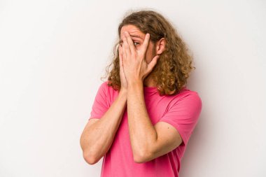 Beyaz arka planda izole edilmiş genç beyaz bir adam parmaklarının arasından korkmuş ve gergin bir şekilde göz kırpıyor..