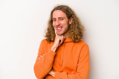 Beyaz arka planda izole olmuş genç beyaz bir adam gülümsüyor ve kendinden emin, çenesine elleriyle dokunuyor..