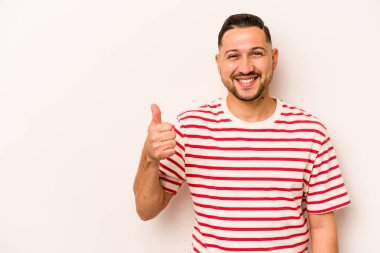 Beyaz arka planda izole edilmiş genç İspanyol adam gülümsüyor ve başparmağını kaldırıyor.