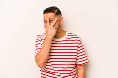 Beyaz arka planda izole edilmiş genç İspanyol adam kameraya parmaklarıyla göz kırpıyor, yüzünü kapatmaktan utanıyor..