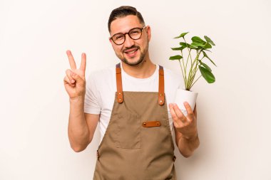 İspanyol bahçıvan beyaz arka planda izole edilmiş bir bitki tutuyor neşeli ve kaygısız parmaklarıyla barış sembolü gösteriyor..