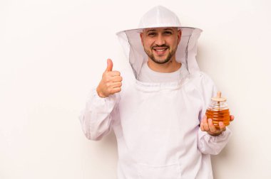 Beyaz arka planda izole edilmiş bal tutan İspanyol arı yetiştiricisi gülümsüyor ve başparmağını kaldırıyor.