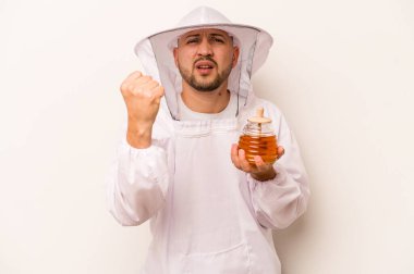 Beyaz arka planda izole edilmiş bal tutan Latin arıcı adam yumruğunu kameraya gösteriyor, agresif yüz ifadesi..