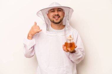 Beyaz arka planda izole edilmiş bal tutan İspanyol arı yetiştiricisi parmaklarıyla bir cep telefonu çağrısı hareketi gösteriyor..