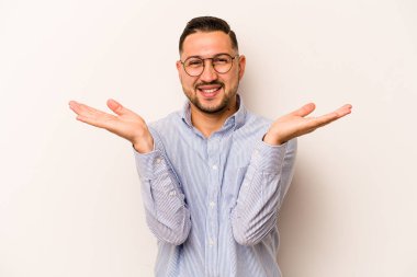 Beyaz arka planda izole edilmiş genç İspanyol adam kollarıyla ölçeği yapıyor, mutlu ve kendinden emin hissediyor..