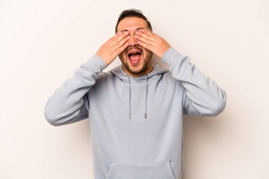 Beyaz arka planda izole edilmiş genç İspanyol adam elleriyle gözlerini kapatıyor, geniş bir gülümsemeyle bir sürpriz bekliyor..