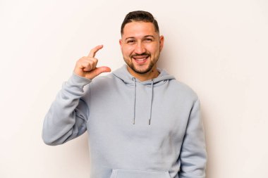 Beyaz arka planda izole edilmiş genç İspanyol adam işaret parmaklarıyla küçük bir şey tutuyor, gülümsüyor ve kendinden emin..