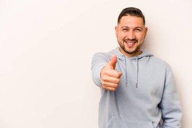 Beyaz arka planda izole edilmiş genç İspanyol adam gülümsüyor ve başparmağını kaldırıyor.