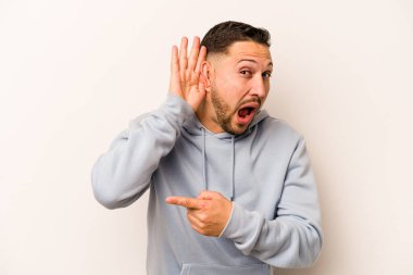Latin kökenli genç bir adam beyaz arka planda bir dedikoduyu dinlemeye çalışıyor..