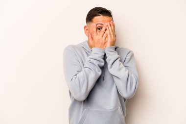 Beyaz arka planda izole edilmiş genç İspanyol adam parmaklarının arasından korkmuş ve gergin bir şekilde göz kırpıyor..