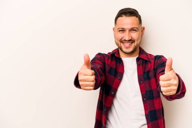 Beyaz arka planda izole edilmiş genç İspanyol adam gülümsüyor ve başparmağını kaldırıyor.