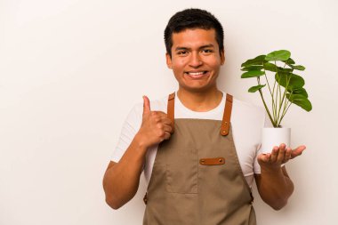 Genç bahçıvan İspanyol bir bitki tutuyor. Beyaz arka planda izole edilmiş. Gülümsüyor ve başparmağını kaldırıyor.