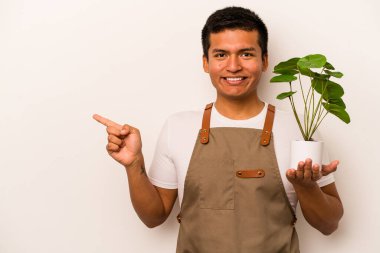 Genç bahçıvan, beyaz arka planda izole edilmiş bir bitki tutuyor gülümsüyor ve boş alanda bir şey gösteriyor..