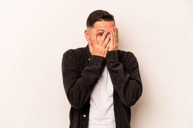 Beyaz arka planda izole edilmiş genç İspanyol adam parmaklarının arasından korkmuş ve gergin bir şekilde göz kırpıyor..