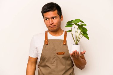Genç bahçıvan İspanyol 'un elinde beyaz arka planda izole edilmiş bir bitki var. Kafası karışmış ve kendinden emin değil..