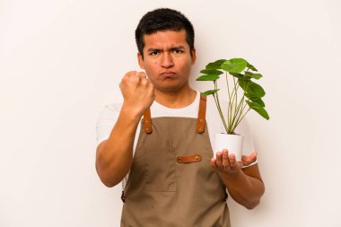 Beyaz arka planda izole edilmiş bir bitki tutan genç bahçıvan kameraya yumruğunu gösteriyor, agresif bir yüz ifadesi..