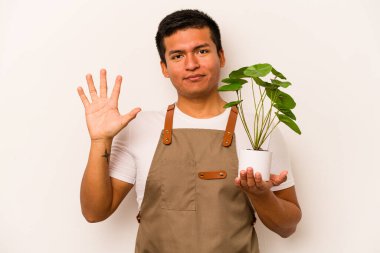 Genç bahçıvan İspanyol bir bitki tutuyor. Beyaz arka planda izole edilmiş. Beş numarayı parmaklarıyla gülümsüyor..