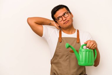 Genç bahçıvan, elinde su dolu bir teneke tutarak beyaz arka planda izole edilmiş kafasının arkasına dokunuyor, düşünüyor ve bir seçim yapıyor..