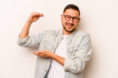 Beyaz arka planda izole edilmiş genç İspanyol adam işaret parmaklarıyla küçük bir şey tutuyor, gülümsüyor ve kendinden emin..