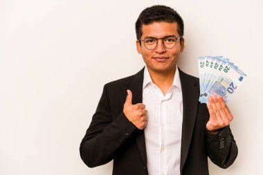 Latin kökenli genç bir iş adamı elinde banknotlar tutarken beyaz arka planda izole edilmiş gülümsüyor ve başparmağını kaldırıyor.