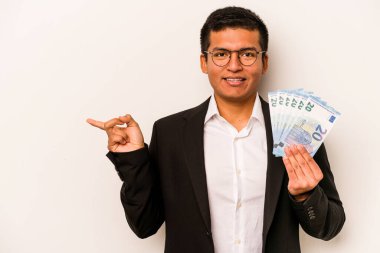 Beyaz arka planda izole edilmiş banknotlar tutan genç İspanyol iş adamı gülümsüyor ve boş alanda bir şeyler gösteriyor..