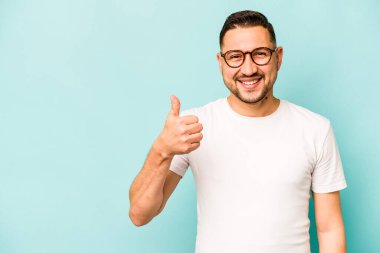 Genç İspanyol adam mavi arka planda izole edilmiş gülümsüyor ve başparmağını kaldırıyor.