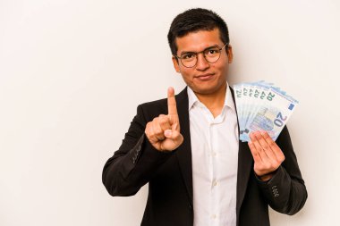 Latin kökenli genç bir iş adamı elinde banknotlar tutarken beyaz arka planda bir numarayı parmağıyla gösteriyor..